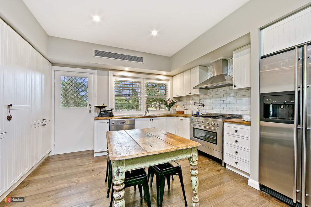Design ideas for a mid-sized country u-shaped separate kitchen in Sydney with a drop-in sink, recessed-panel cabinets, white cabinets, wood benchtops, white splashback, porcelain splashback, stainless steel appliances, dark hardwood floors and with island.