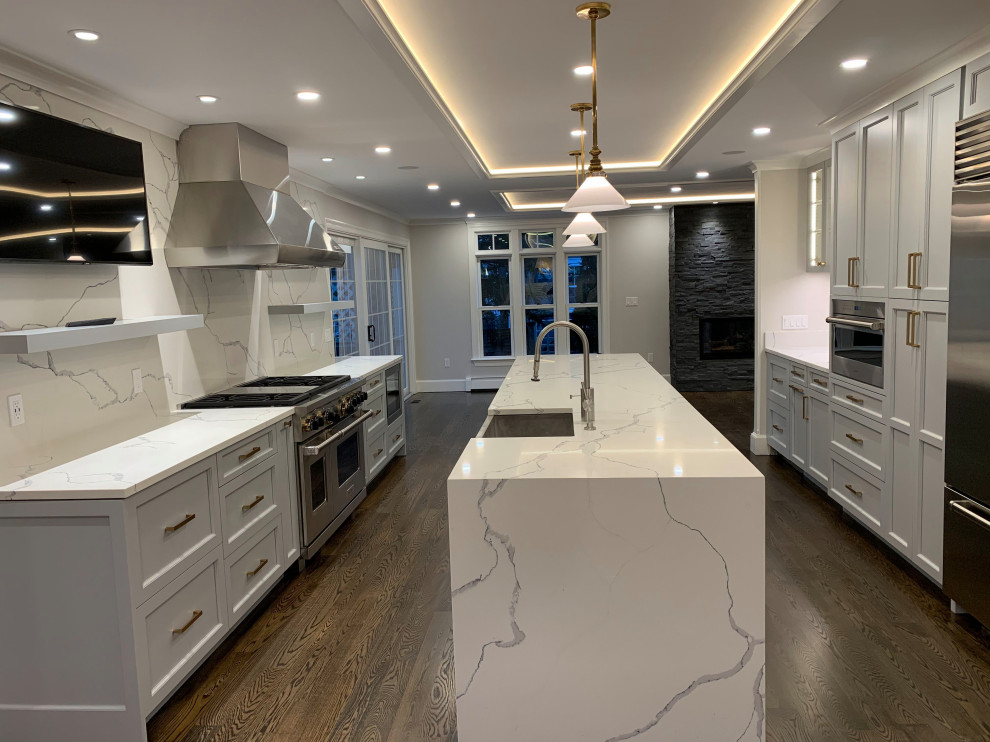 Large transitional galley dark wood floor and brown floor enclosed kitchen photo in Boston with a farmhouse sink, raised-panel cabinets, quartzite countertops, white backsplash, stone slab backsplash, stainless steel appliances, an island, white countertops and gray cabinets