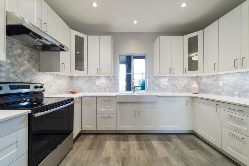 This is an example of a mid-sized contemporary u-shaped separate kitchen in Vancouver with a farmhouse sink, shaker cabinets, white cabinets, quartz benchtops, grey splashback, marble splashback, stainless steel appliances, laminate floors, no island, brown floor and white benchtop.