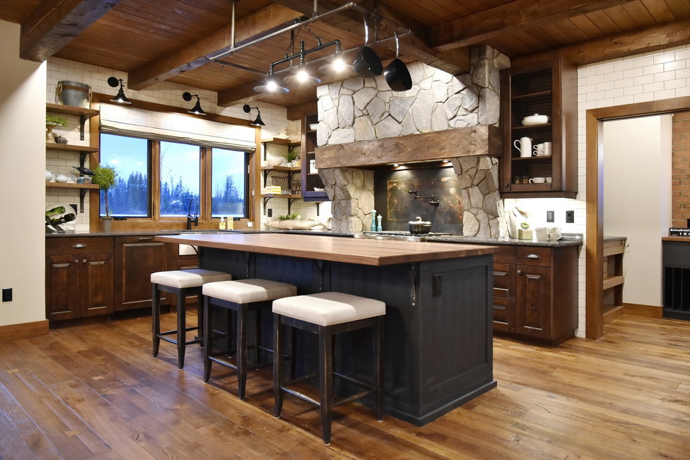 This is an example of a mid-sized country l-shaped open plan kitchen in Calgary with raised-panel cabinets, dark wood cabinets, wood benchtops, medium hardwood floors, with island, a farmhouse sink, white splashback, subway tile splashback, stainless steel appliances, brown floor and brown benchtop.