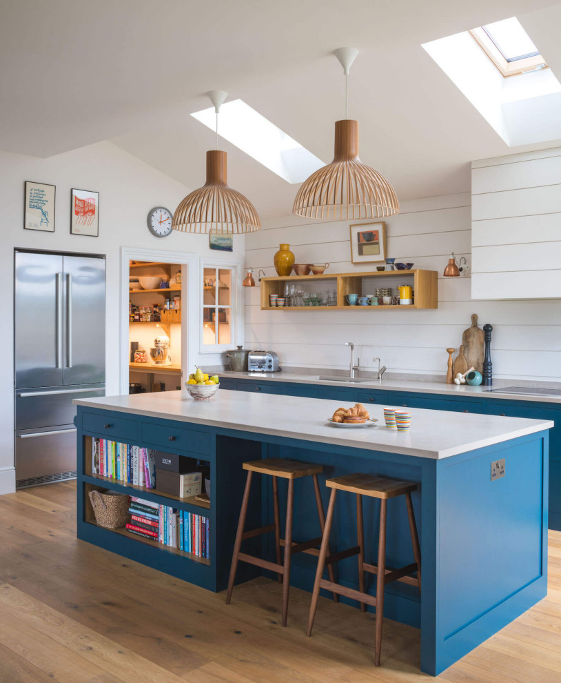Mid-sized transitional open plan kitchen in Other with a drop-in sink, shaker cabinets, blue cabinets, quartzite benchtops, light hardwood floors, with island, white benchtop, white splashback, stainless steel appliances and brown floor.
