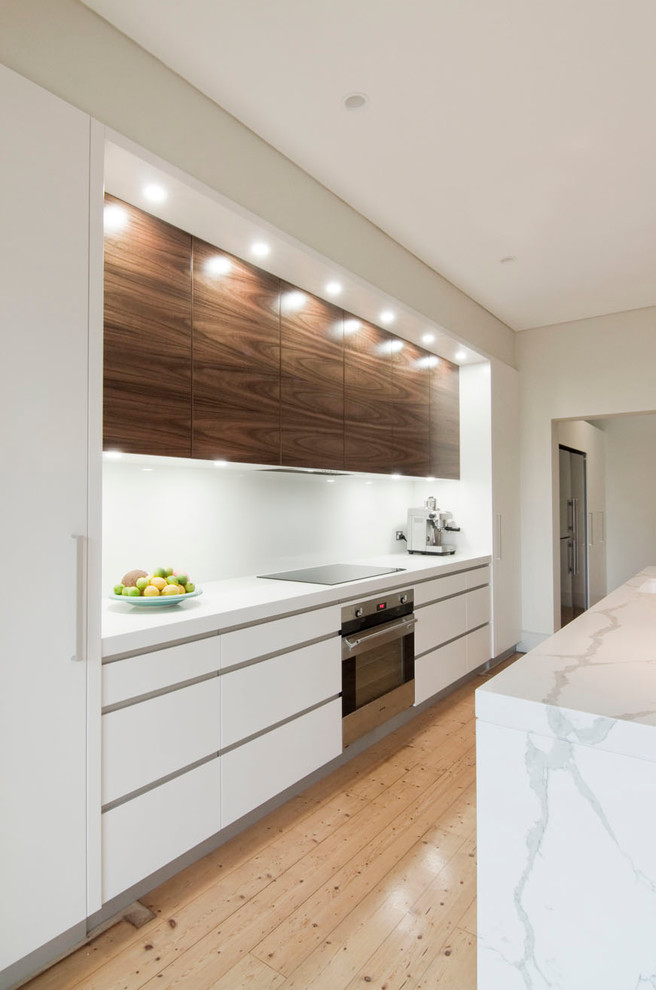 Photo of a large modern single-wall open plan kitchen in Sydney with an undermount sink, flat-panel cabinets, quartz benchtops, white splashback, glass sheet splashback, stainless steel appliances, light hardwood floors, with island and medium wood cabinets.