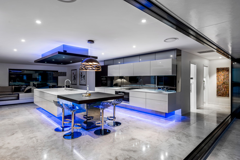 This is an example of an expansive contemporary galley open plan kitchen in Brisbane with an undermount sink, glass-front cabinets, beige cabinets, marble benchtops, black splashback, mirror splashback, black appliances, travertine floors, with island and beige floor.
