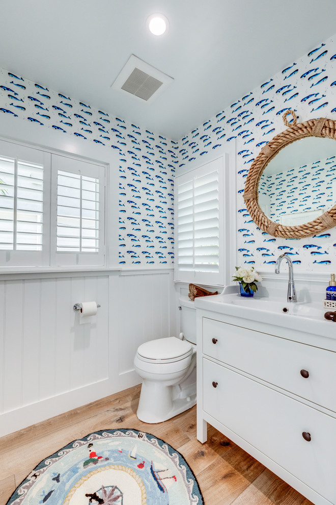 Small beach style powder room in Los Angeles with furniture-like cabinets, white cabinets, multi-coloured walls, light hardwood floors, brown floor and white benchtops.