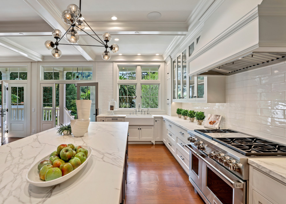 Photo of a medium sized traditional grey and white u-shaped open plan kitchen in San Francisco with a belfast sink, shaker cabinets, white cabinets, marble worktops, white splashback, metro tiled splashback, stainless steel appliances, medium hardwood flooring, an island, brown floors, white worktops and a coffered ceiling.