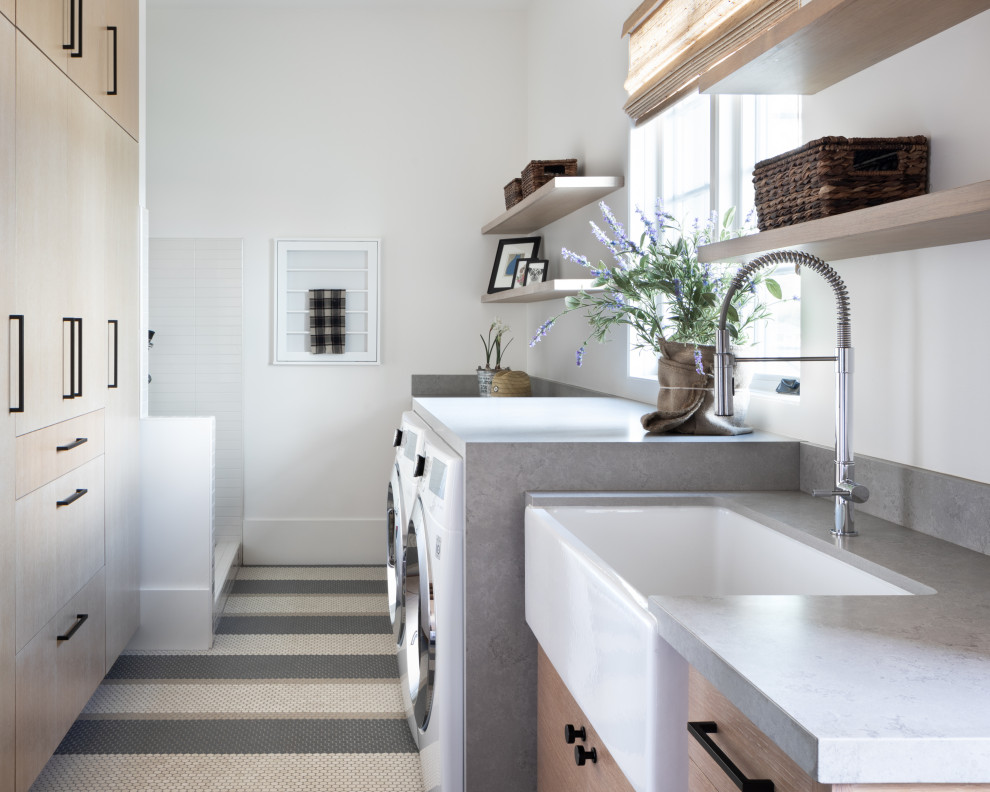 Country galley dedicated laundry room in San Francisco with a farmhouse sink, flat-panel cabinets, light wood cabinets, quartz benchtops, white walls, porcelain floors, a side-by-side washer and dryer and grey benchtop.