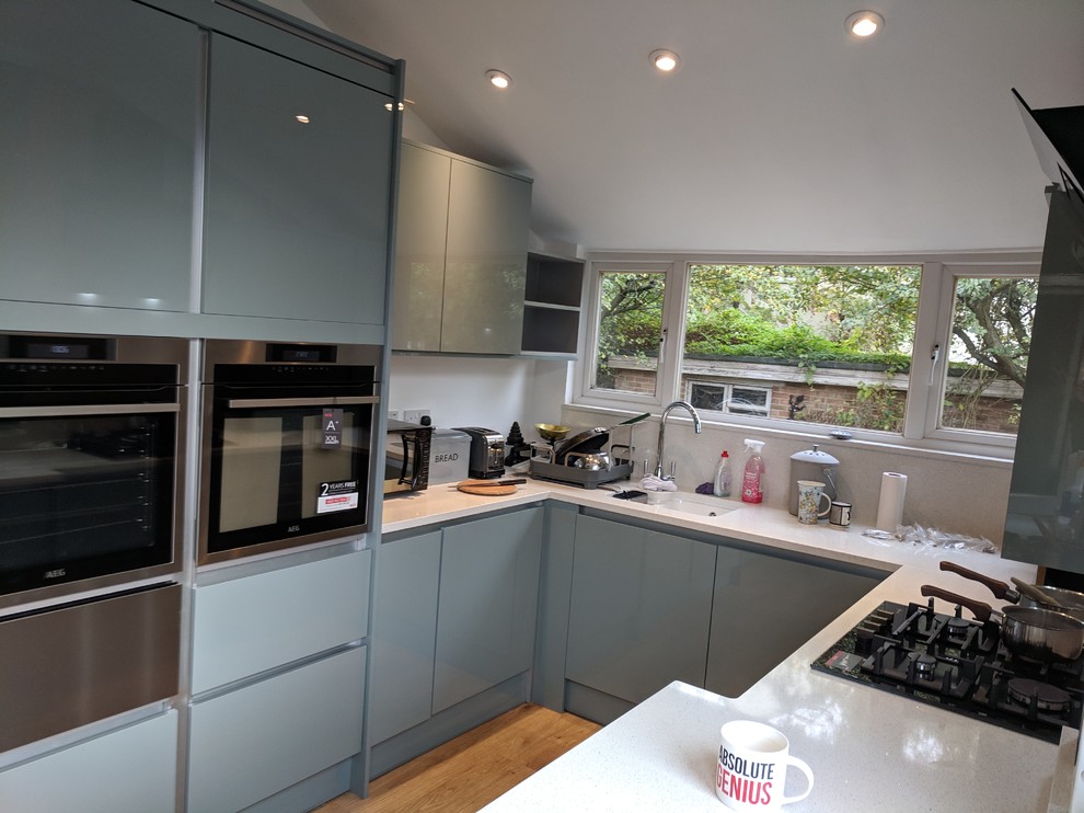 Design ideas for a mid-sized modern u-shaped separate kitchen in London with an undermount sink, flat-panel cabinets, turquoise cabinets, quartzite benchtops, white splashback, stone slab splashback, black appliances, vinyl floors, no island, brown floor and white benchtop.