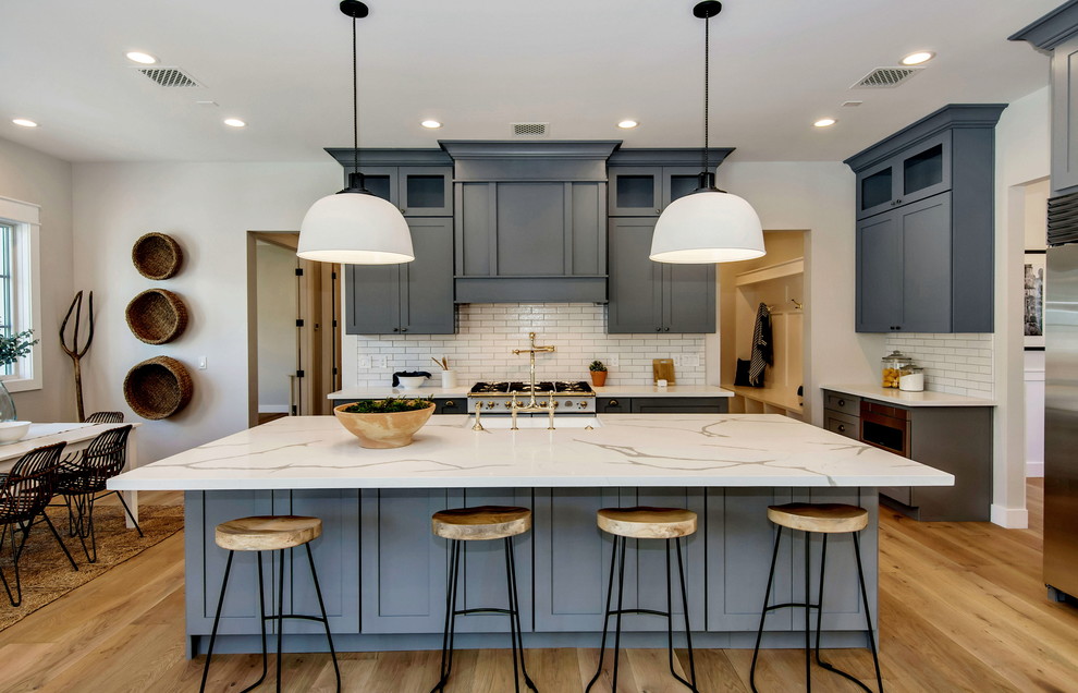 Photo of a large country l-shaped open plan kitchen in Phoenix with a farmhouse sink, shaker cabinets, grey cabinets, quartzite benchtops, white splashback, subway tile splashback, stainless steel appliances, light hardwood floors, with island, brown floor and white benchtop.