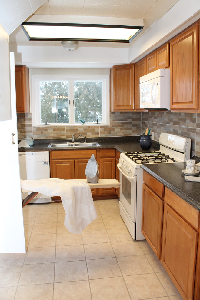 Photo of a small country l-shaped separate kitchen in New York with a double-bowl sink, raised-panel cabinets, medium wood cabinets, solid surface benchtops, beige splashback, ceramic splashback, white appliances and ceramic floors.