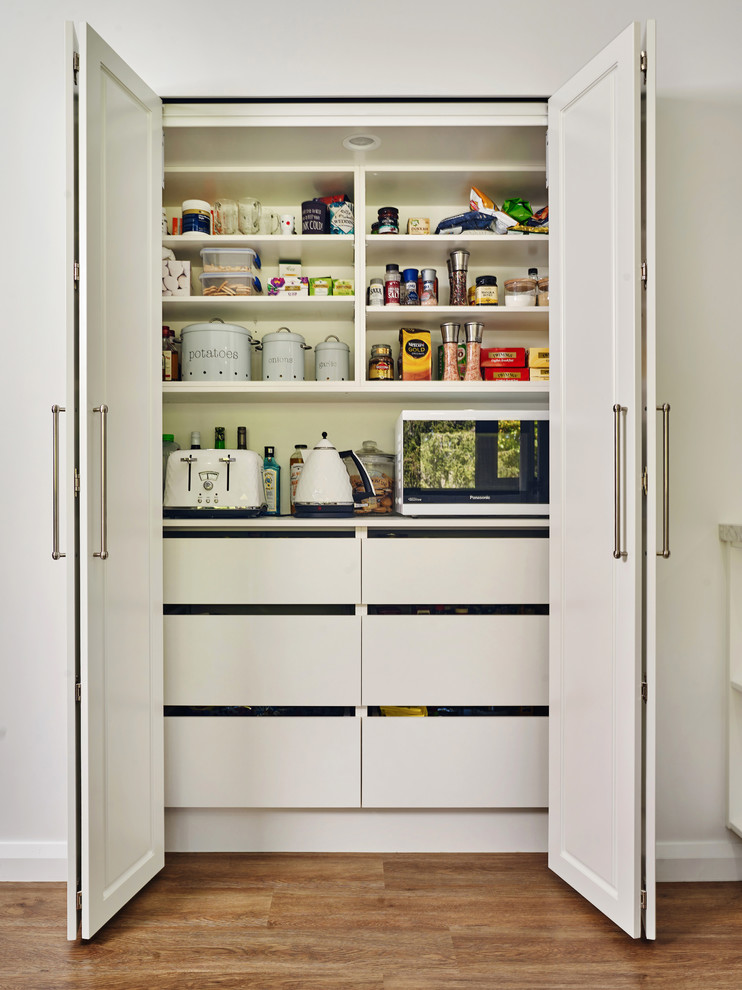 This is an example of a mid-sized u-shaped kitchen pantry with a drop-in sink, raised-panel cabinets, white cabinets, quartz benchtops, grey splashback, ceramic splashback, black appliances, light hardwood floors, with island, brown floor and white benchtop.