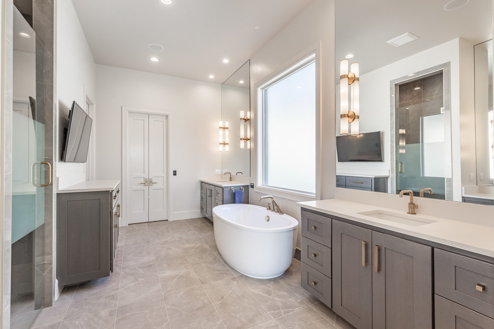 This is an example of a transitional master bathroom in Oklahoma City with shaker cabinets, grey cabinets, a freestanding tub, an alcove shower, white walls, an undermount sink, beige floor and a hinged shower door.