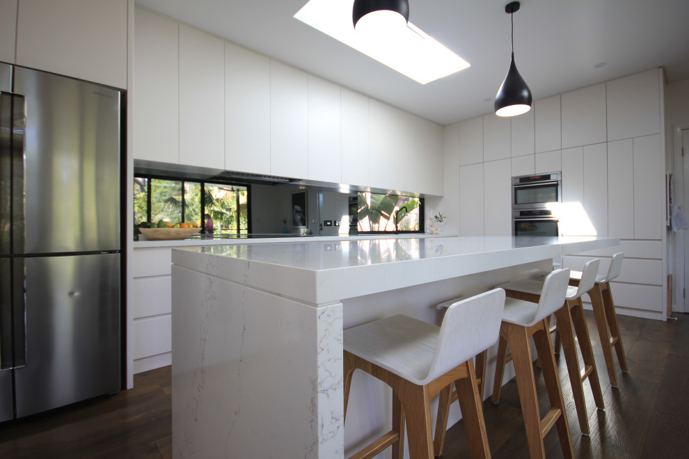 This is an example of a mid-sized modern l-shaped open plan kitchen in Sydney with an undermount sink, white cabinets, quartz benchtops, glass sheet splashback, stainless steel appliances, with island, brown floor and white benchtop.