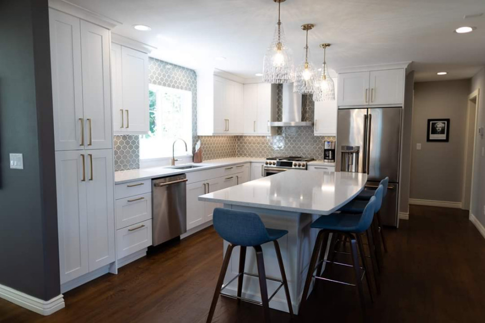 Photo of a medium sized classic l-shaped kitchen/diner in Seattle with a submerged sink, shaker cabinets, white cabinets, engineered stone countertops, grey splashback, glass tiled splashback, stainless steel appliances, dark hardwood flooring, an island, brown floors and white worktops.