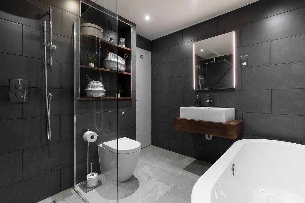 This is an example of a large modern bathroom in London with raised-panel cabinets, dark wood cabinets, a freestanding tub, a one-piece toilet, gray tile, cement tile, grey walls, ceramic floors, a pedestal sink, wood benchtops, an open shower, an open shower and brown benchtops.