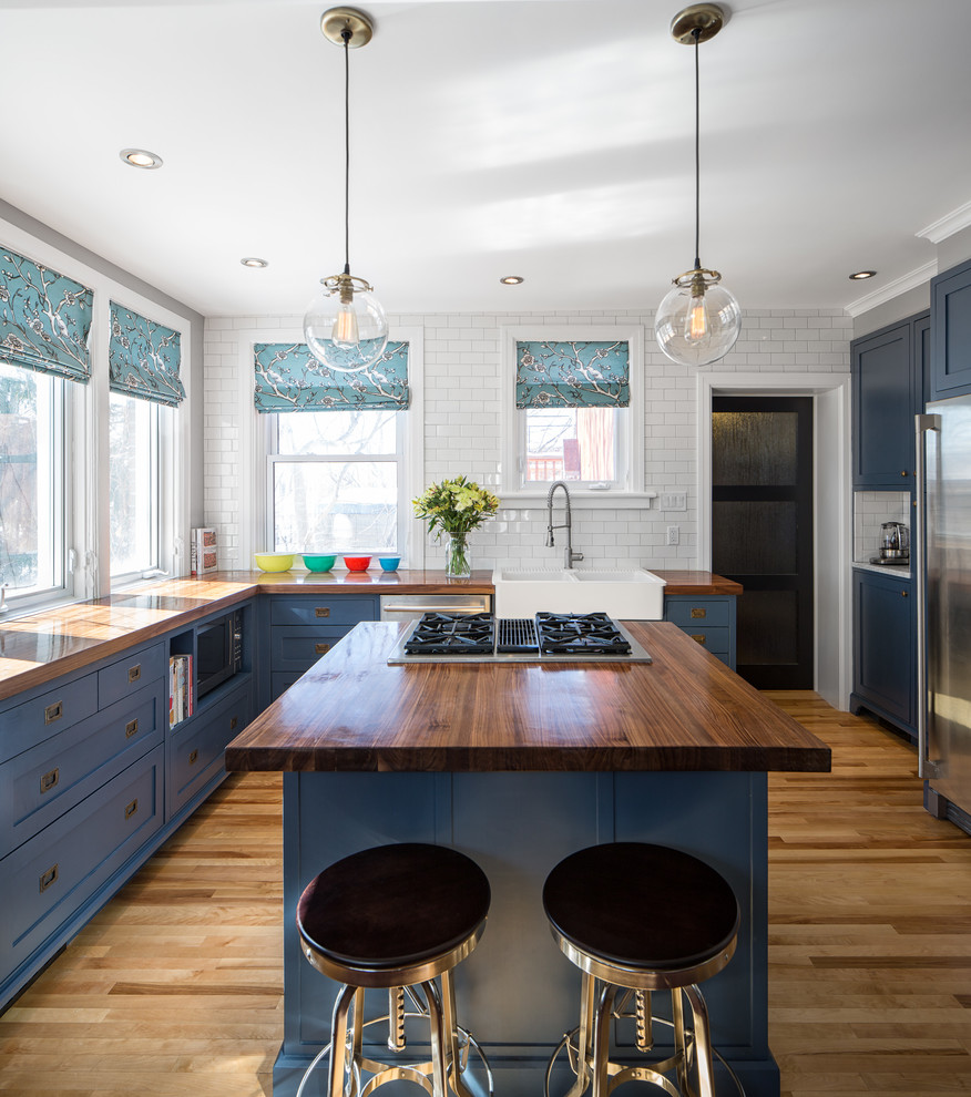 Photo of a transitional l-shaped kitchen in Ottawa with a farmhouse sink, recessed-panel cabinets, wood benchtops, white splashback, subway tile splashback, stainless steel appliances, medium hardwood floors, with island and blue cabinets.