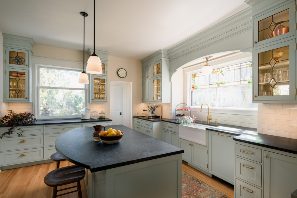 This is an example of a small traditional u-shaped kitchen in Seattle with a farmhouse sink, beaded inset cabinets, green cabinets, soapstone benchtops, white splashback, subway tile splashback, panelled appliances, with island, black benchtop, medium hardwood floors and brown floor.
