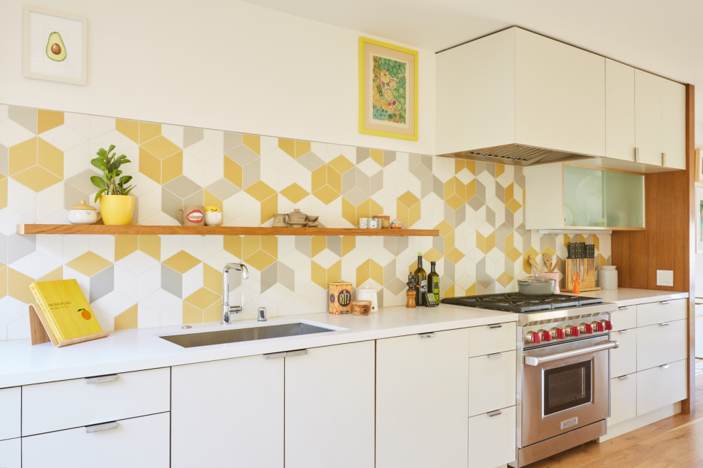 This is an example of a midcentury l-shaped eat-in kitchen in Los Angeles with a single-bowl sink, flat-panel cabinets, white cabinets, quartz benchtops, yellow splashback, ceramic splashback, stainless steel appliances, light hardwood floors, a peninsula, brown floor and white benchtop.