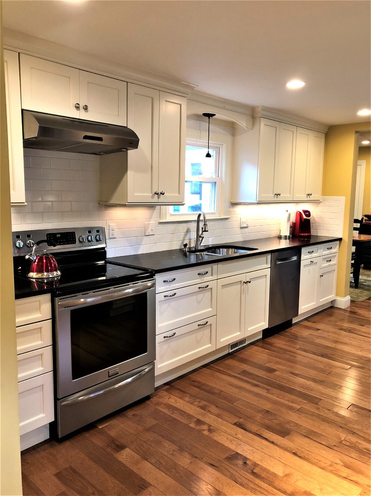 Inspiration for a small transitional galley medium tone wood floor and brown floor enclosed kitchen remodel in Boston with an undermount sink, shaker cabinets, white cabinets, granite countertops, white backsplash, subway tile backsplash, stainless steel appliances and black countertops