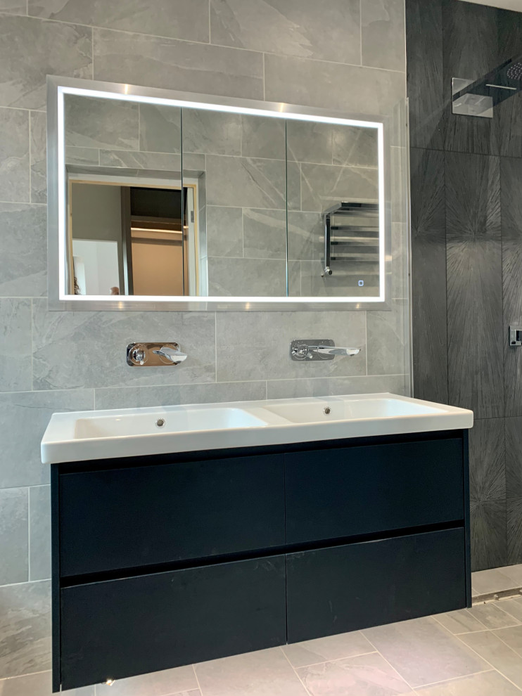 Photo of a medium sized ensuite wet room bathroom in Hertfordshire with flat-panel cabinets, blue cabinets, a wall mounted toilet, grey tiles, porcelain tiles, grey walls, porcelain flooring, a wall-mounted sink, grey floors, an open shower, a wall niche, double sinks, a floating vanity unit and a coffered ceiling.