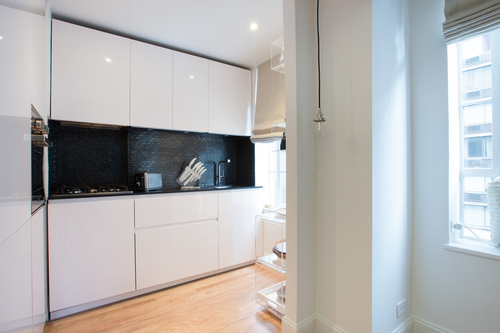 Photo of a contemporary kitchen in New York with flat-panel cabinets, black splashback and white cabinets.