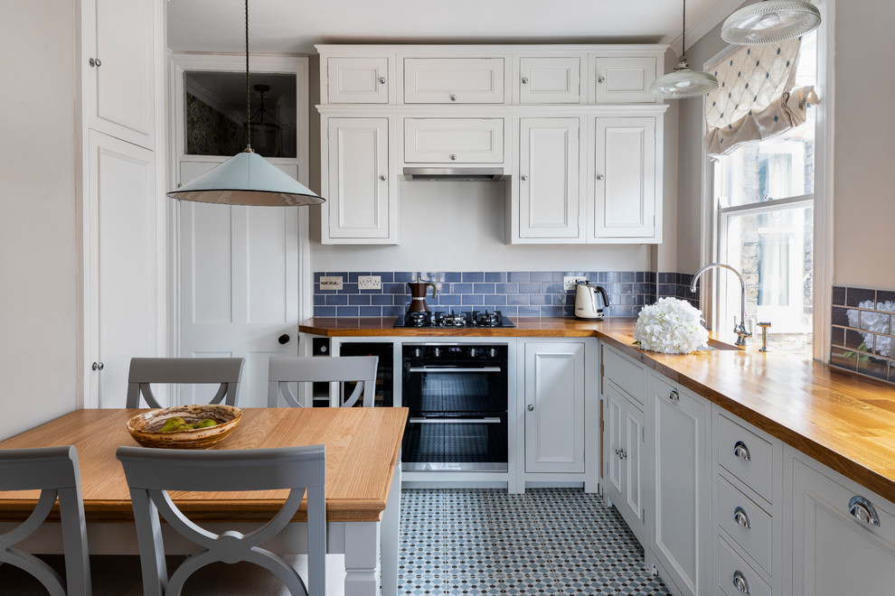 Design ideas for a mid-sized traditional l-shaped eat-in kitchen in London with a farmhouse sink, white cabinets, wood benchtops, blue splashback, ceramic splashback, panelled appliances, ceramic floors, no island, blue floor, recessed-panel cabinets and brown benchtop.
