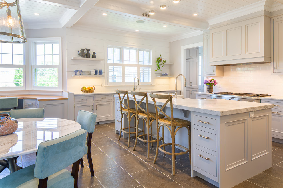 This is an example of a large beach style u-shaped eat-in kitchen in New York with a farmhouse sink, flat-panel cabinets, grey cabinets, marble benchtops, white splashback, ceramic splashback, stainless steel appliances, limestone floors, with island, grey floor and white benchtop.