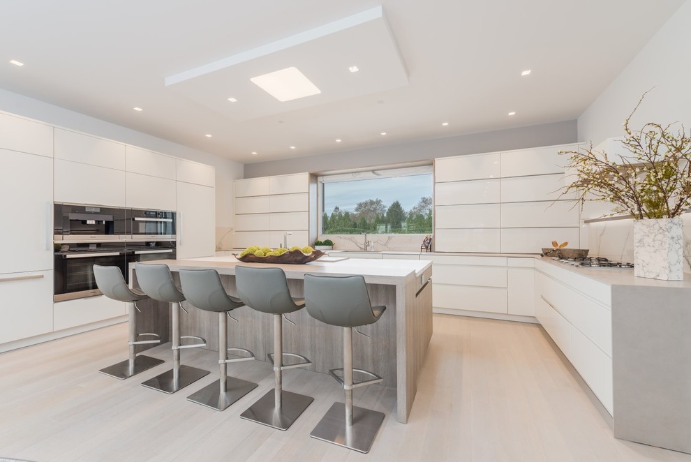 Design ideas for a contemporary u-shaped kitchen in New York with flat-panel cabinets, white cabinets, grey splashback, black appliances, light hardwood floors, with island, beige floor and grey benchtop.