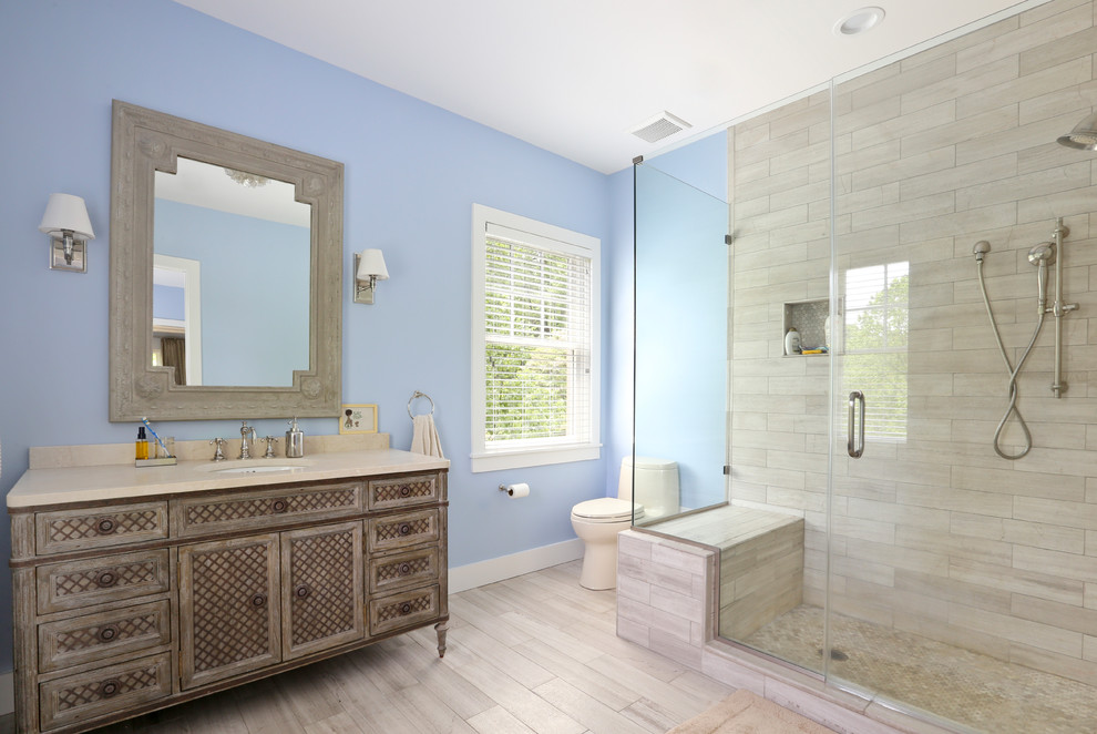 This is an example of a mid-sized country master bathroom in DC Metro with furniture-like cabinets, an alcove shower, a one-piece toilet, gray tile, porcelain tile, blue walls, an undermount sink, engineered quartz benchtops, a hinged shower door, brown cabinets, vinyl floors and beige floor.
