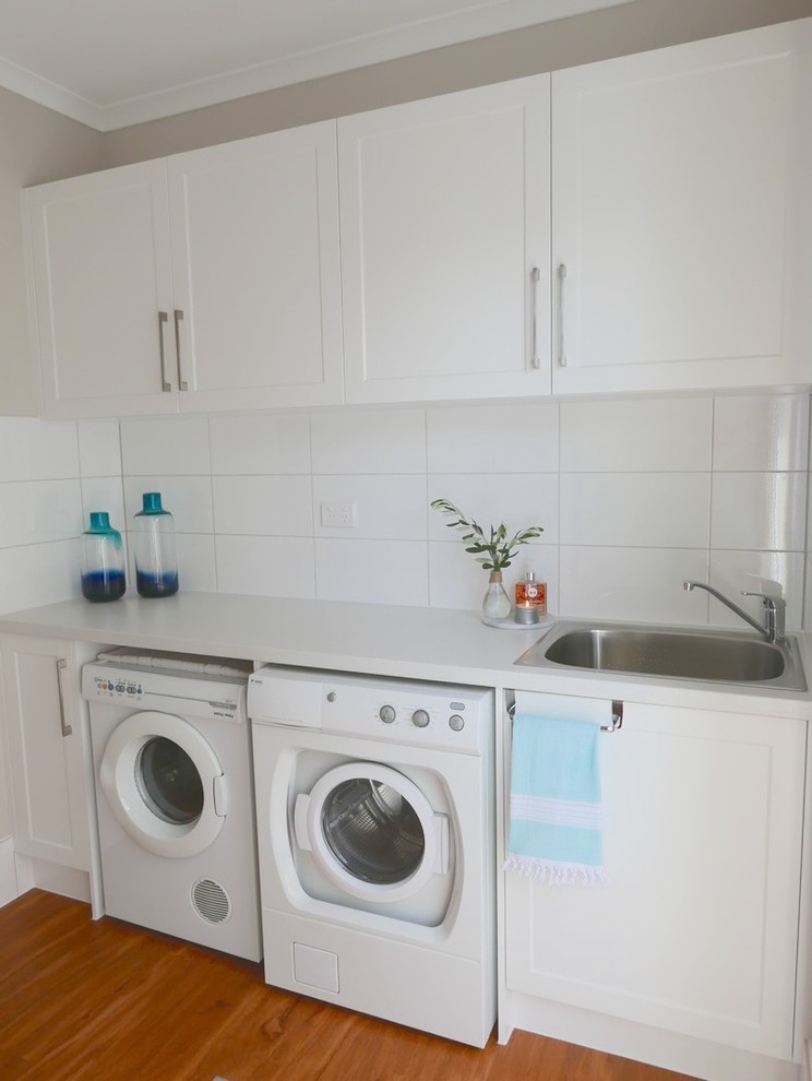 Photo of a small beach style galley dedicated laundry room in Brisbane with a drop-in sink, shaker cabinets, white cabinets, quartz benchtops, beige walls, light hardwood floors and a side-by-side washer and dryer.