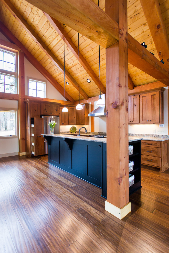 Large mountain style l-shaped medium tone wood floor open concept kitchen photo with a farmhouse sink, shaker cabinets, medium tone wood cabinets, granite countertops, stainless steel appliances and an island