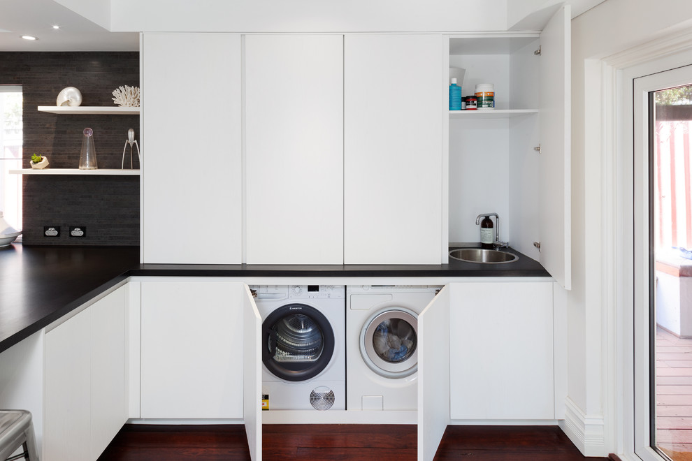 Modern laundry room in Perth with a single-bowl sink, white cabinets, granite benchtops, white walls, dark hardwood floors, black benchtop and a concealed washer and dryer.