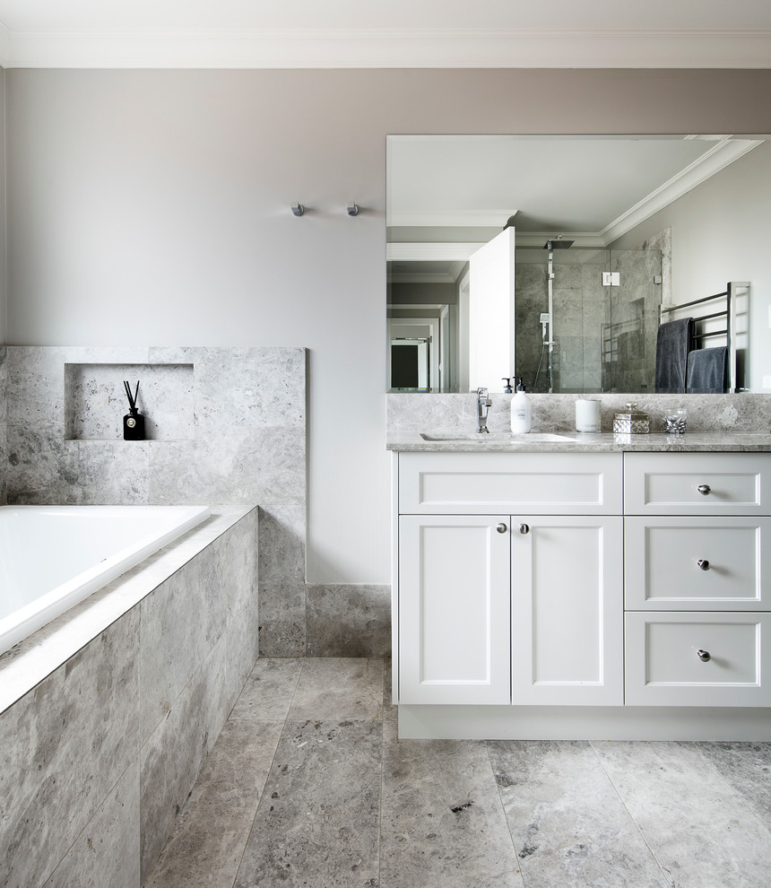 Photo of a transitional bathroom in Sydney with shaker cabinets, white cabinets, a drop-in tub, a one-piece toilet, gray tile, stone slab, grey walls, limestone floors, an undermount sink and limestone benchtops.