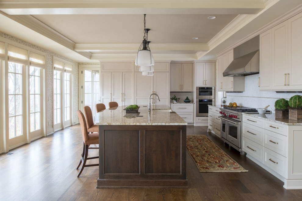 Large transitional l-shaped eat-in kitchen in Minneapolis with shaker cabinets, beige cabinets, with island, a farmhouse sink, granite benchtops, white splashback, mosaic tile splashback, stainless steel appliances, dark hardwood floors, brown floor, multi-coloured benchtop and exposed beam.