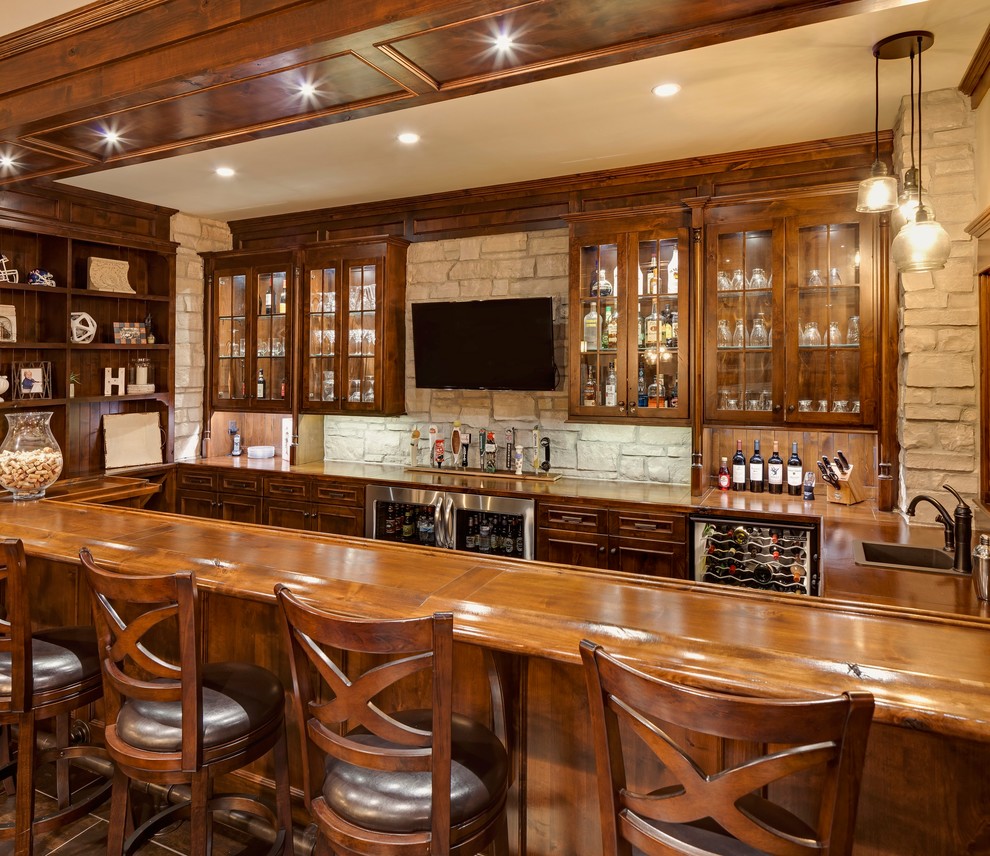 This is an example of a large country u-shaped seated home bar in Chicago with a drop-in sink, glass-front cabinets, dark wood cabinets, wood benchtops, grey splashback, stone tile splashback, porcelain floors, brown floor and brown benchtop.