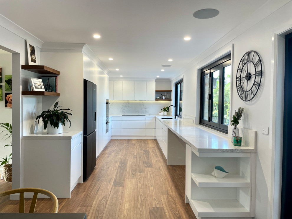 This is an example of a large u-shaped eat-in kitchen in Sydney with an undermount sink, flat-panel cabinets, white cabinets, quartz benchtops, white splashback, engineered quartz splashback, black appliances, medium hardwood floors, brown floor and white benchtop.