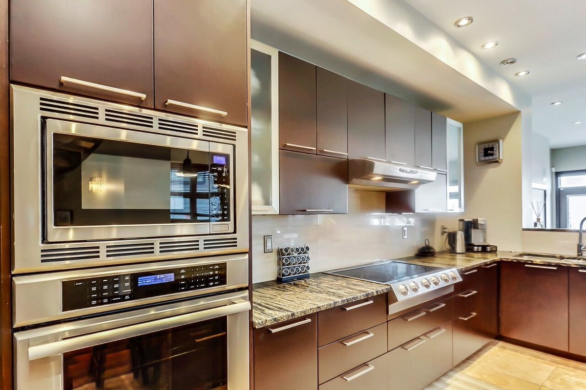 Large transitional l-shaped travertine floor open concept kitchen photo in Calgary with a double-bowl sink, flat-panel cabinets, dark wood cabinets, granite countertops, beige backsplash, stainless steel appliances and an island