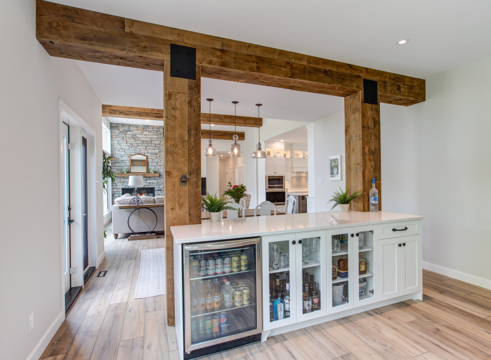 Photo of a large country single-wall home bar in Other with shaker cabinets, white cabinets, quartz benchtops, medium hardwood floors, brown floor and beige benchtop.