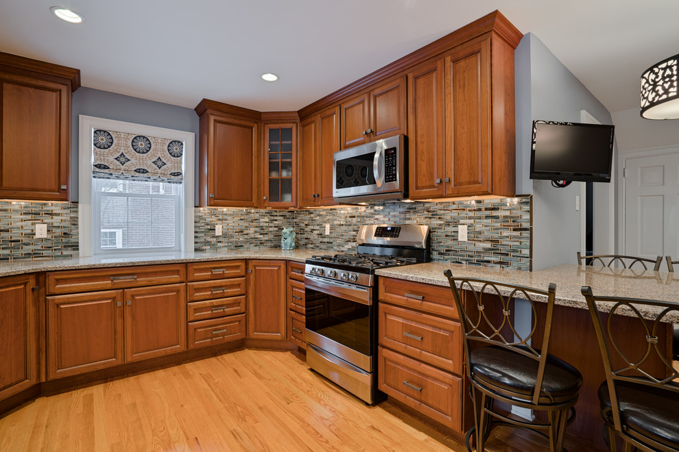 This is an example of a mid-sized traditional u-shaped eat-in kitchen in Philadelphia with a farmhouse sink, raised-panel cabinets, medium wood cabinets, granite benchtops, multi-coloured splashback, glass tile splashback, stainless steel appliances, light hardwood floors, a peninsula and brown floor.