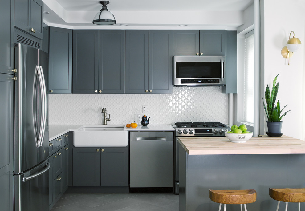 Inspiration for a mid-sized country l-shaped eat-in kitchen in New York with a farmhouse sink, quartz benchtops, white splashback, stainless steel appliances, porcelain floors, shaker cabinets, grey cabinets and a peninsula.
