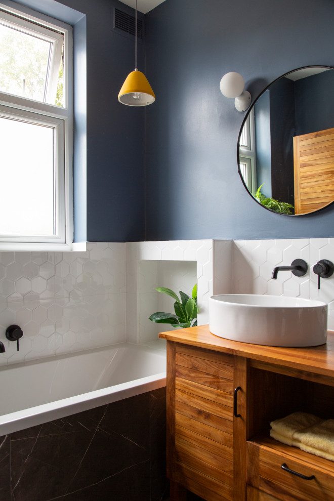 Photo of a small eclectic kids wet room bathroom in London with louvered cabinets, brown cabinets, a drop-in tub, a wall-mount toilet, white tile, porcelain tile, blue walls, porcelain floors, wood benchtops, black floor, brown benchtops, a single vanity, a freestanding vanity and wood.
