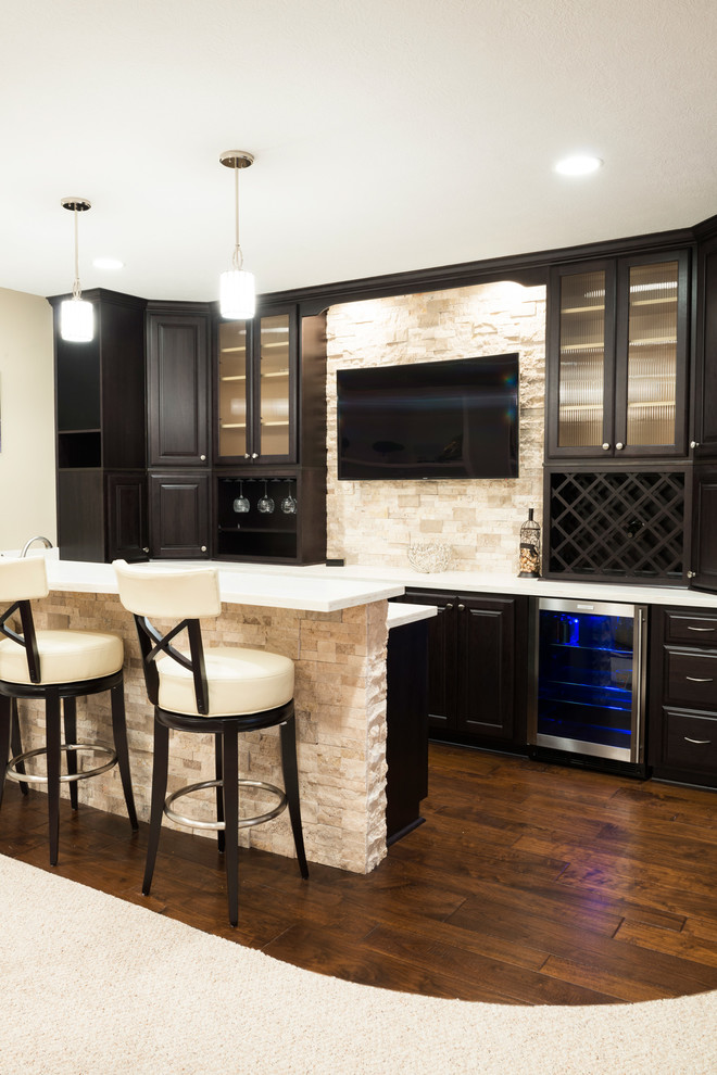 Design ideas for a mid-sized contemporary galley seated home bar in Indianapolis with dark wood cabinets, solid surface benchtops, beige splashback, stone tile splashback, dark hardwood floors and raised-panel cabinets.