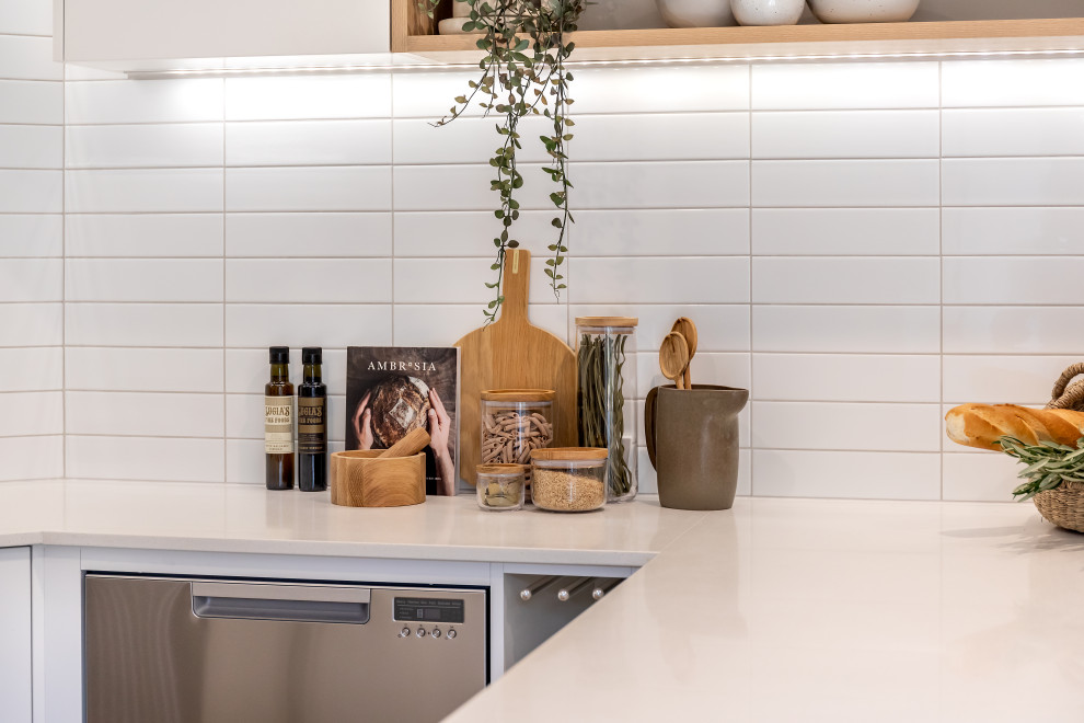 This is an example of a small contemporary u-shaped kitchen/diner in Adelaide with a submerged sink, flat-panel cabinets, white cabinets, engineered stone countertops, white splashback, metro tiled splashback, stainless steel appliances, laminate floors, a breakfast bar, brown floors and white worktops.