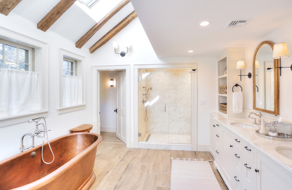 This is an example of a large country master bathroom in Bridgeport with shaker cabinets, white cabinets, a freestanding tub, an alcove shower, a one-piece toilet, white tile, porcelain tile, white walls, light hardwood floors, an undermount sink, engineered quartz benchtops, beige floor, a hinged shower door and white benchtops.
