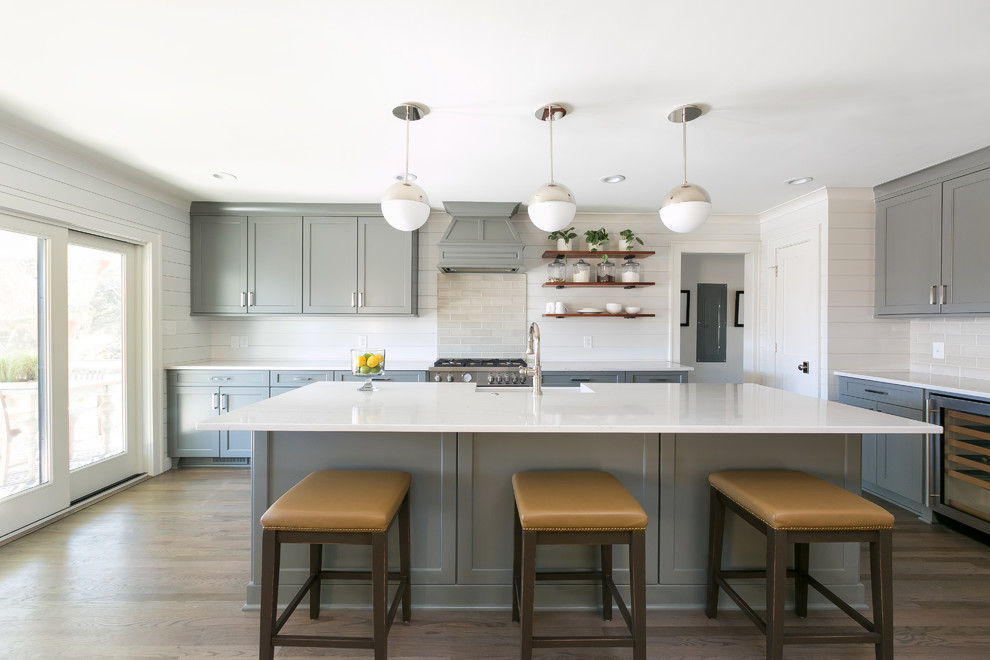 Mid-sized modern l-shaped open plan kitchen with a farmhouse sink, shaker cabinets, grey cabinets, beige splashback, subway tile splashback, stainless steel appliances, medium hardwood floors, with island, brown floor and white benchtop.