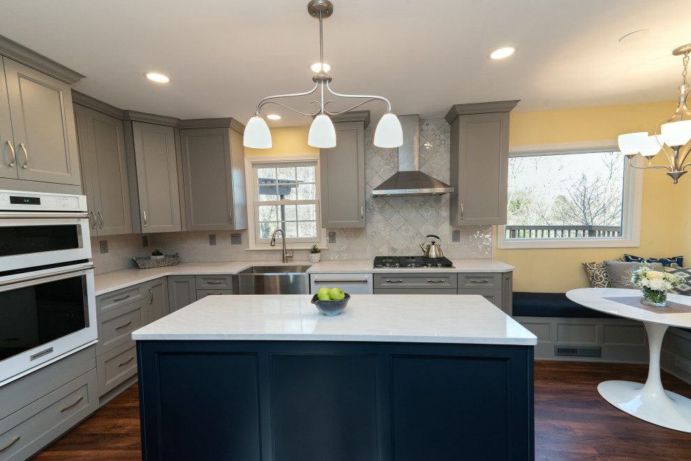 This is an example of a large transitional u-shaped eat-in kitchen in Baltimore with a farmhouse sink, recessed-panel cabinets, grey cabinets, quartzite benchtops, white splashback, subway tile splashback, white appliances, dark hardwood floors, with island, brown floor and white benchtop.