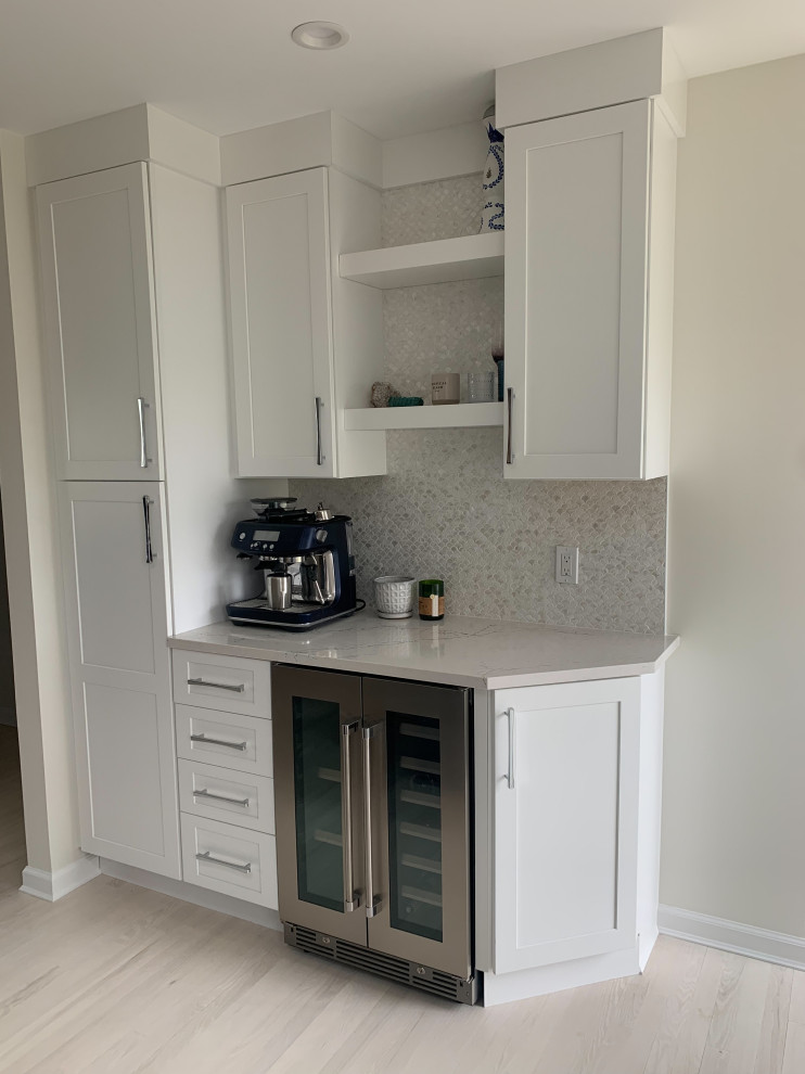 Photo of a small beach style single-wall home bar in New York with no sink, shaker cabinets, white cabinets, quartz benchtops, multi-coloured splashback, mosaic tile splashback, light hardwood floors, white floor and white benchtop.