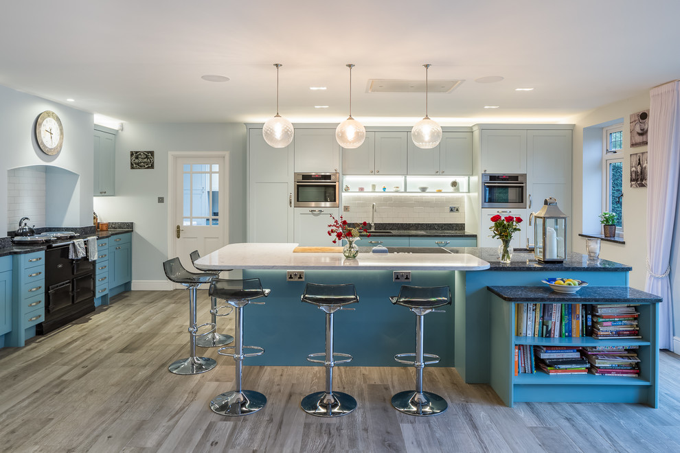 Design ideas for a large transitional u-shaped kitchen in Hertfordshire with a drop-in sink, shaker cabinets, blue cabinets, granite benchtops, white splashback, stone slab splashback, stainless steel appliances, medium hardwood floors, black benchtop, with island and grey floor.