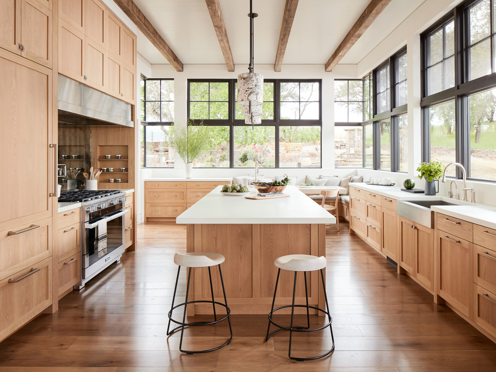 This is an example of a country u-shaped eat-in kitchen in San Francisco with a farmhouse sink, shaker cabinets, medium wood cabinets, stainless steel appliances, medium hardwood floors, with island, brown floor and white benchtop.