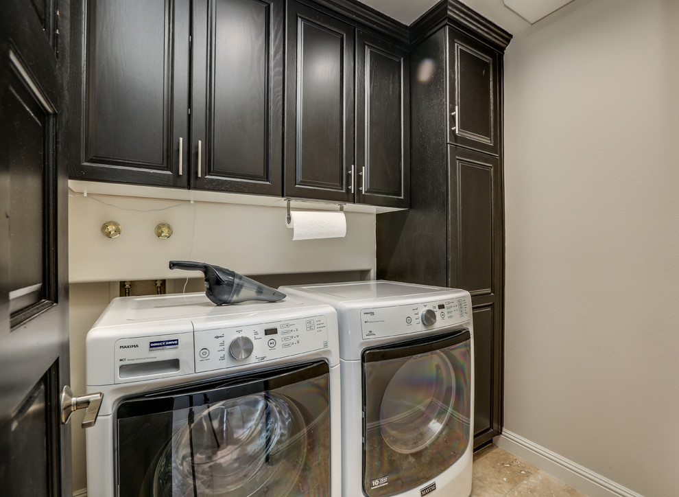 Inspiration for a mid-sized tropical single-wall dedicated laundry room in Other with raised-panel cabinets, black cabinets, grey walls, travertine floors, a side-by-side washer and dryer, brown floor and grey benchtop.