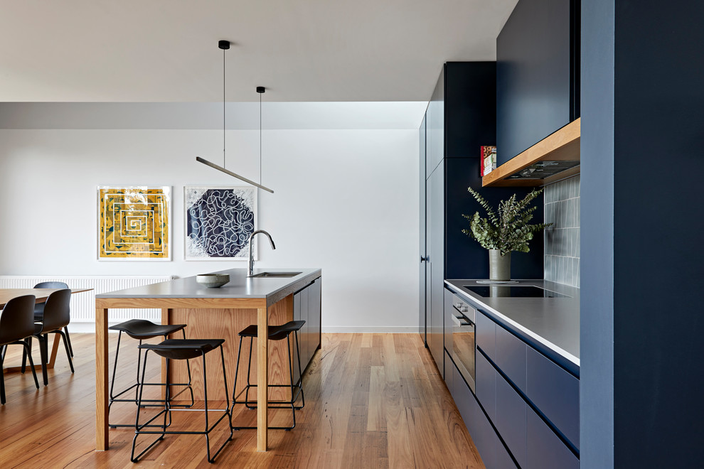 Photo of a large modern galley eat-in kitchen in Melbourne with a double-bowl sink, flat-panel cabinets, blue cabinets, quartz benchtops, grey splashback, ceramic splashback, black appliances, with island, grey benchtop, medium hardwood floors and brown floor.